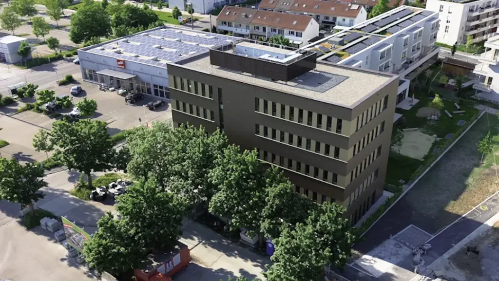 Luftaufnahme des Gebäudes des Therapie Zentrums Augsburg, umgeben von Bäumen und benachbarten Gebäuden, mit einem großen Parkplatz und einer Solaranlage auf dem angrenzenden Gebäude.